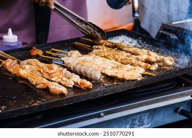 Closeup of a hand grilling assorted skewers of marinated meat, sausages, and vegetables on a hot griddle. Smoke and spices enhance the street food vibe. - Powered by Shutterstock