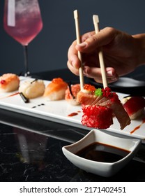 Close-up Hand Holding Bamboo Chopsticks A Piece Of Sushi While Soaking It In Soy Sauce At Restaurant. Front View. Japanese Cuisine Concept.