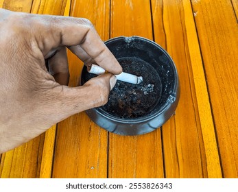 Close-up of a hand holding a cigarette above a black ashtray on a polished wooden surface - Powered by Shutterstock