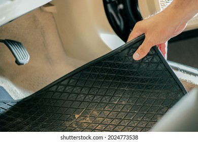 Close-up Of A Hand Holding A Dirty Black Rubber Car Floor Mat With Accelerator And Brake Inside The Cabin Before Dusting, Vacuuming, Dry Cleaning, Washing The Car.