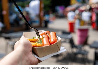 Close-up of hand holding a disposable plate with grilled sausages and roasted potatoes, in an outdoor setting with blurred people in background. Perfect for illustrating outdoor dining or food events. - Powered by Shutterstock