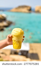 Close-up of a hand holding fresh pineapple slices in a plastic cup. The background shows the blurred turquoise water of the Blue Lagoon in Gozo, Malta on a sunny day.