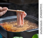 A close-up of a hand holding a fresh slice of meat with chopsticks, ready for cooking in a bubbling hot pot broth, highlighting the culinary preparation.