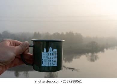 Close-up of hand holding green metal mug with cityscape print, set against serene foggy lake and forest backdrop on misty morning. Travel, relaxation, and enjoying nature quiet moments.  - Powered by Shutterstock