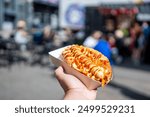 close-up of a hand holding a hot dog with various toppings in paper tray, set against blurred street fair background. vibrant scene captures essence of outdoor food festivals and street food culture.