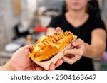 A close-up of a hand holding a hot dog with mustard and ketchup, with an out-of-focus person in the background. The hot dog is the main focus, showcasing its toppings and the casual dining experience.