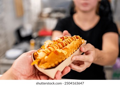 A close-up of a hand holding a hot dog with mustard and ketchup, with an out-of-focus person in the background. The hot dog is the main focus, showcasing its toppings and the casual dining experience. - Powered by Shutterstock