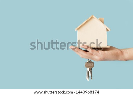 Close-up of hand holding keys and wooden house model against blue background