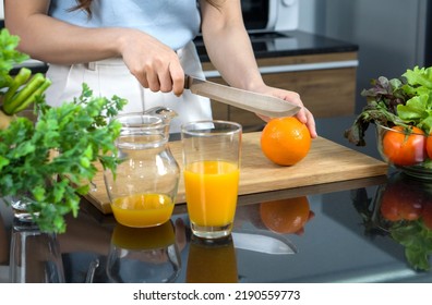 Closeup Hand Holding Knife Cutting Orange Fruit On A Wooden Chop Board. Jar With Mixed Fruit Juice  And Glass Bowl With A Variety Of Fruits And Vegetables Is Placed On The Kitchen Counter.