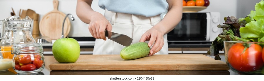 Closeup Hand Holding Knife Cutting Green Cucumber On A Wooden Chop Board (front View). A Glass Bowl With Various Kind Of Vegetable, Tomato Jar And Fruit Juice Are On Kitchen Counter.