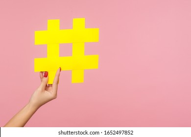 Closeup of hand holding large yellow paper hashtag symbol next to copy space, hash sign of famous media content, social media marketing and blog promotion. studio shot isolated on pink background