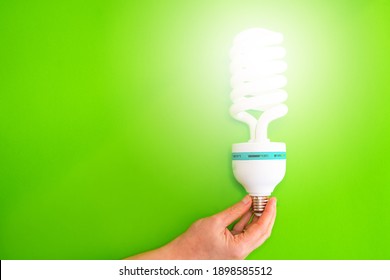 Close-up Of A Hand Holding A Lit Led Bulb Against A Green Background. Concept Of Energy Saving At Home. Copy Space