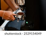 A close-up of a hand holding a portability filled with coffee grounds, positioned near an espresso machine. The machine features a sleek design with a green indicator log