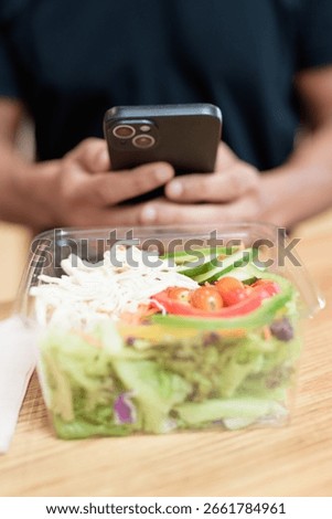 Close-up of a hand holding smartphone while interacting with fresh salad in takeaway container. Shot indoors in mall food court. Healthy eating, mobile lifestyle, and social media concept.