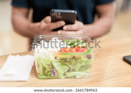 Close-up of a hand holding smartphone while interacting with fresh salad in takeaway container. Shot indoors in mall food court. Healthy eating, mobile lifestyle, and social media concept.
