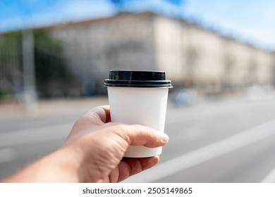 Close-up of a hand holding a white takeaway coffee cup with a black lid, against a blurred urban background. Ideal for themes of coffee culture, urban lifestyle, and on-the-go beverages. - Powered by Shutterstock