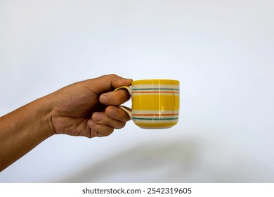 Close-up of a hand holding a yellow ceramic mug with a colorful striped pattern against a plain white background. The vibrant yellow mug features red, green, and white stripes. - Powered by Shutterstock