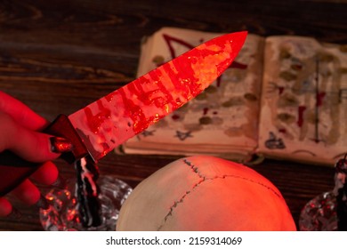Close-up Hand Holds Knife Stained With Blood On The Satanic Book Background. Antichrist Satanic Ritual Concept.