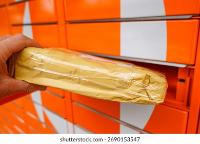 Close-up of a hand inserting a wrapped package into a bright orange parcel locker, representing shipping, delivery, online shopping, and e-commerce logistics