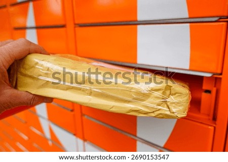 Close-up of a hand inserting a wrapped package into a bright orange parcel locker, representing shipping, delivery, online shopping, and e-commerce logistics