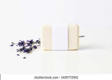 Closeup of hand made herbal soap bar in blank paper label package and bunch of lavender flowers on white table backround. Spa concept. Skin product mockup scene. Cosmetic product.