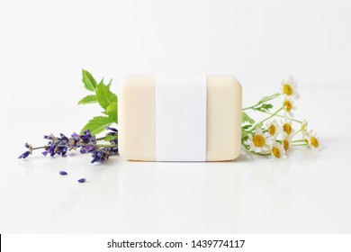 Closeup of hand made herbal soap bar in blank paper label package. Mint leaves, lavender and feverfew flowers on white table backround. Spa concept. Skin product mockup scene. Cosmetic product.