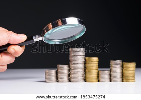 Closeup hand with magnifying glass analyze on money heap coins that growing unstable, uncertain income and unpredictable finance, or finding money resources concept