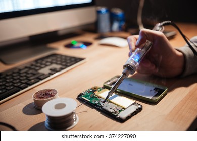 Closeup of hand of man sitting near computer and repairing cell phone with soldering iron