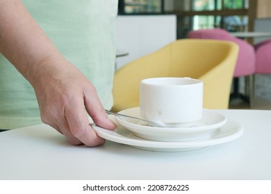 Close-up Of A Hand Picking Up A Stack Of Plates And A Dirty Coffee Cup From The Table. The Waiter In The Cafe Cleans The Dirty Dishes From The Table. Cafeteria Maintenance Work.