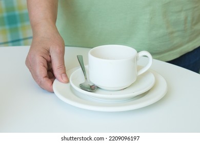 Close-up Of A Hand Picking Up A Stack Of Plates And A Dirty Coffee Cup From The Table. The Waiter In The Cafe Cleans The Dirty Dishes From The Table. Cafeteria Maintenance Work.