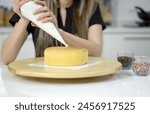 Closeup hand piping white cream on a sponge cake, creating a decorative pattern on the top of a round, light-colored cake that