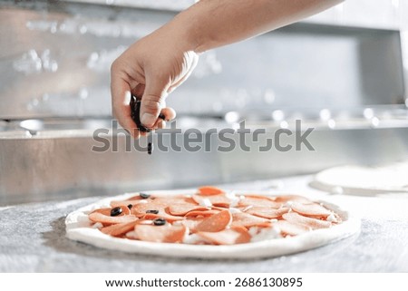 Closeup of hand pizzamaker preparing pepperoni pizza with olives in pizzeria kitchen.