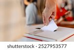 Close-up of hand placing ballot into voting box at election. Ballot and voting box symbolize democracy and elections. Hand placing ballot in election voting box, casting a vote at election