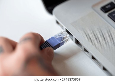 Close-up of a hand plugging an Ethernet cable into a laptop port, symbolizing connectivity and technology. The hand has visible tattoos.