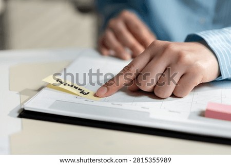 Close-up of a hand pointing to a sticky note on a planner during business scheduling, illustrating task management, project planning, productivity, organization, deadline tracking and time management.