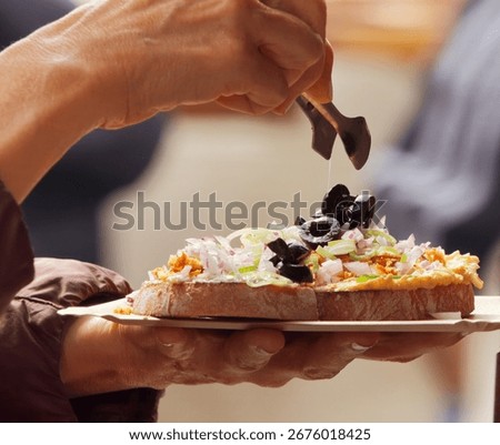 Close-up of hand preparing slice of bread with spread, onions, and black olives at Naplavka farmers market in Prague, Czech Republic.
