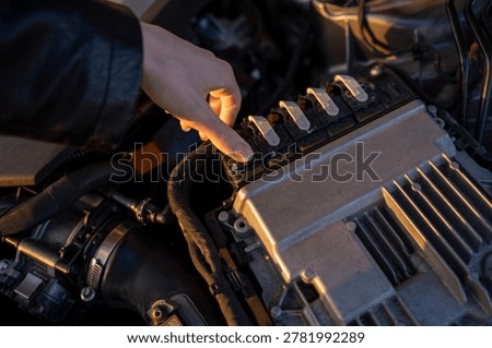Close-up of hand pressing the locking tab on an electrical connector in a car engine compartment, showing vehicle wiring and electronic components.