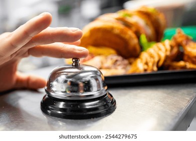 A close-up of a hand pressing a service bell on a counter, with a blurred background featuring a sandwich and fries. This image captures the concept of prompt service in the food industry. - Powered by Shutterstock