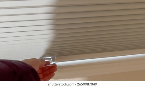 A close-up of a hand pulling down the white blinds on a basement window. The interior wall and windowsill are softly illuminated.