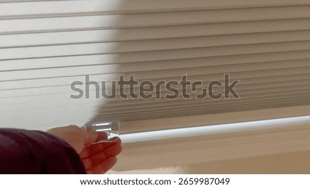 A close-up of a hand pulling down the white blinds on a basement window. The interior wall and windowsill are softly illuminated.
