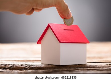 Closeup of hand putting coin in house piggy bank on wood