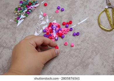 Close-up of a hand selecting a single red bead, with a pile of pink and purple pony beads and gold scissors nearby. Concept of DIY jewelry making, hobby, creative pastime, and artistic craft projects.