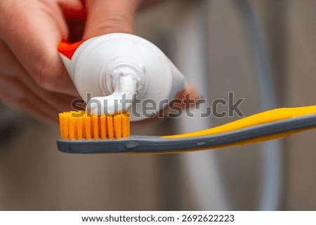 Close-up of hand squeezing white toothpaste from red and white tube onto bright orange toothbrush bristles with water droplets, symbolizing oral hygiene, freshness, dental care, and morning routine