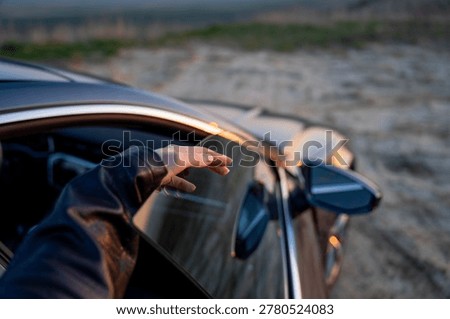 Close-up of hand touching the upper edge of a partially lowered side window on an open modern car door outdoors in evening light.