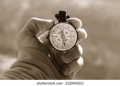 Close-up Hand Of A Tourist With An Authentic Compass On The Background Of A Mountain Road Landscape In Warm Sepia Toning