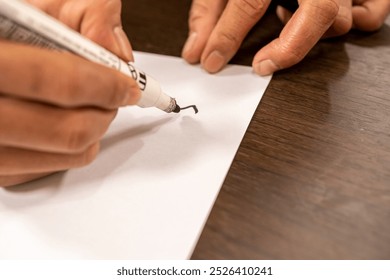 Close-up of a hand using a black marker to write on a blank white paper, showing creative expression or note-taking on a wooden surface. - Powered by Shutterstock