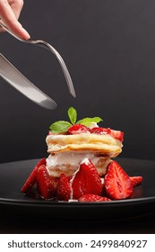 Close-up of hand using knife and fork to cut into a stack of fluffy pancakes topped with fresh strawberries and whipped cream.