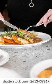  A close-up of a hand using a knife and fork to cut into a plate of crispy chicken, accompanied by vegetables and garnished with fresh herbs, served on a textured table.
