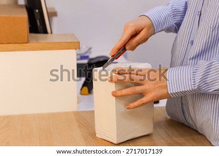 Close-up of hand using knife to open sealed white parcel on desk at order pickup station.