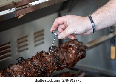 Close-up of a hand using a meat thermometer to check the temperature of grilled meat on a rotisserie for perfect cooking.